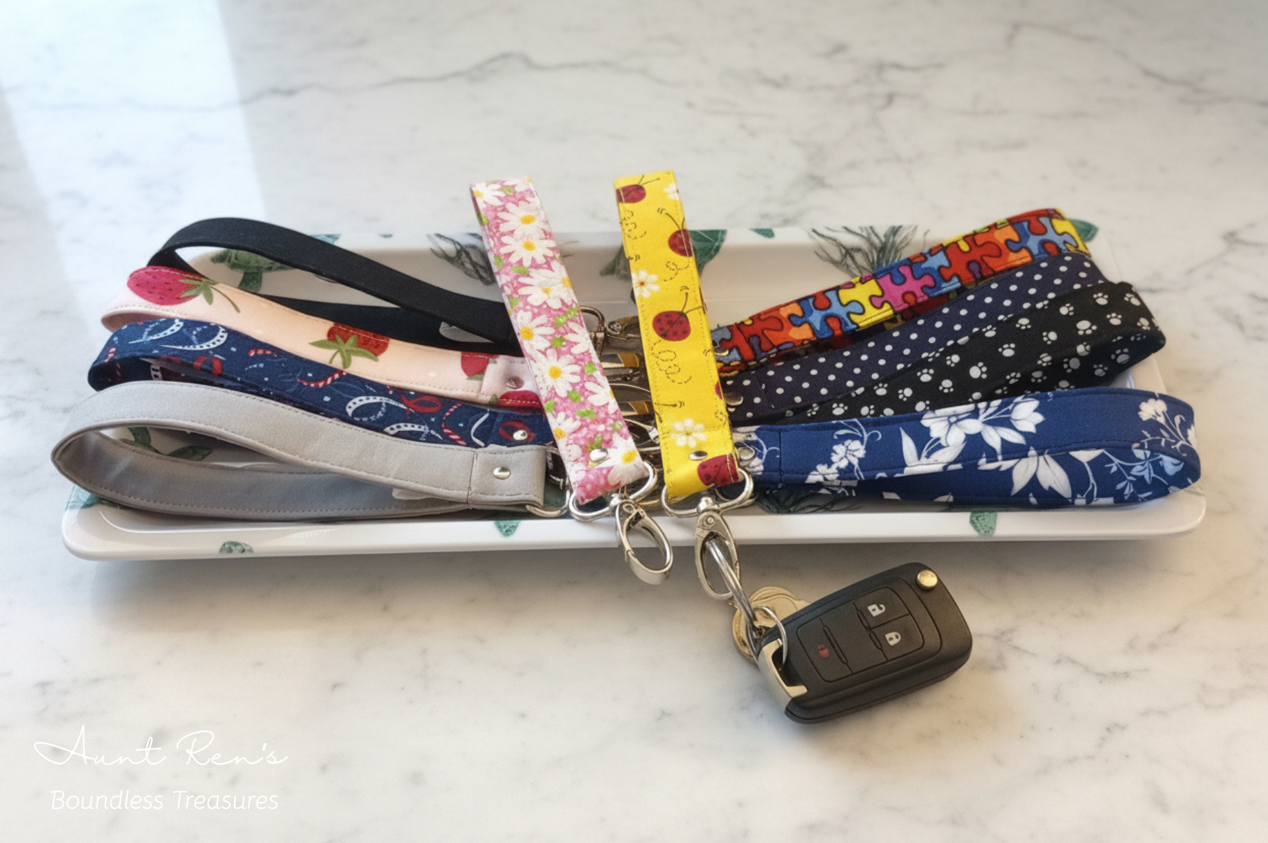 Ten fabric wristlet keychains in various colorful patterns, displayed on a white dish over a white marble surface. One wristlet is attached to a black car key fob.