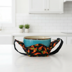 A ceramic bowl with a turquoise blue glazed exterior and a rustic brown rim, paired with a black fabric bowl holder, printed with a red chili pepper pattern, shown against a plain white background.