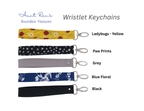 Four fabric wristlet keychains with metal keyrings are shown on a white background, each labeled with a design name and color: Ladybugs - Yellow, Paw Prints, Grey, and Blue Floral, plus a black option. The brand name Aunt Ren's Boundless Treasures appears at the top left.