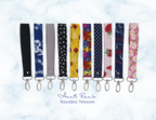 Ten fabric wristlets with silver metal keychains are lined up horizontally on a white and light blue marble background. The wristlets feature various patterns including stripes, polka dots, floral prints, and colorful retro motifs.